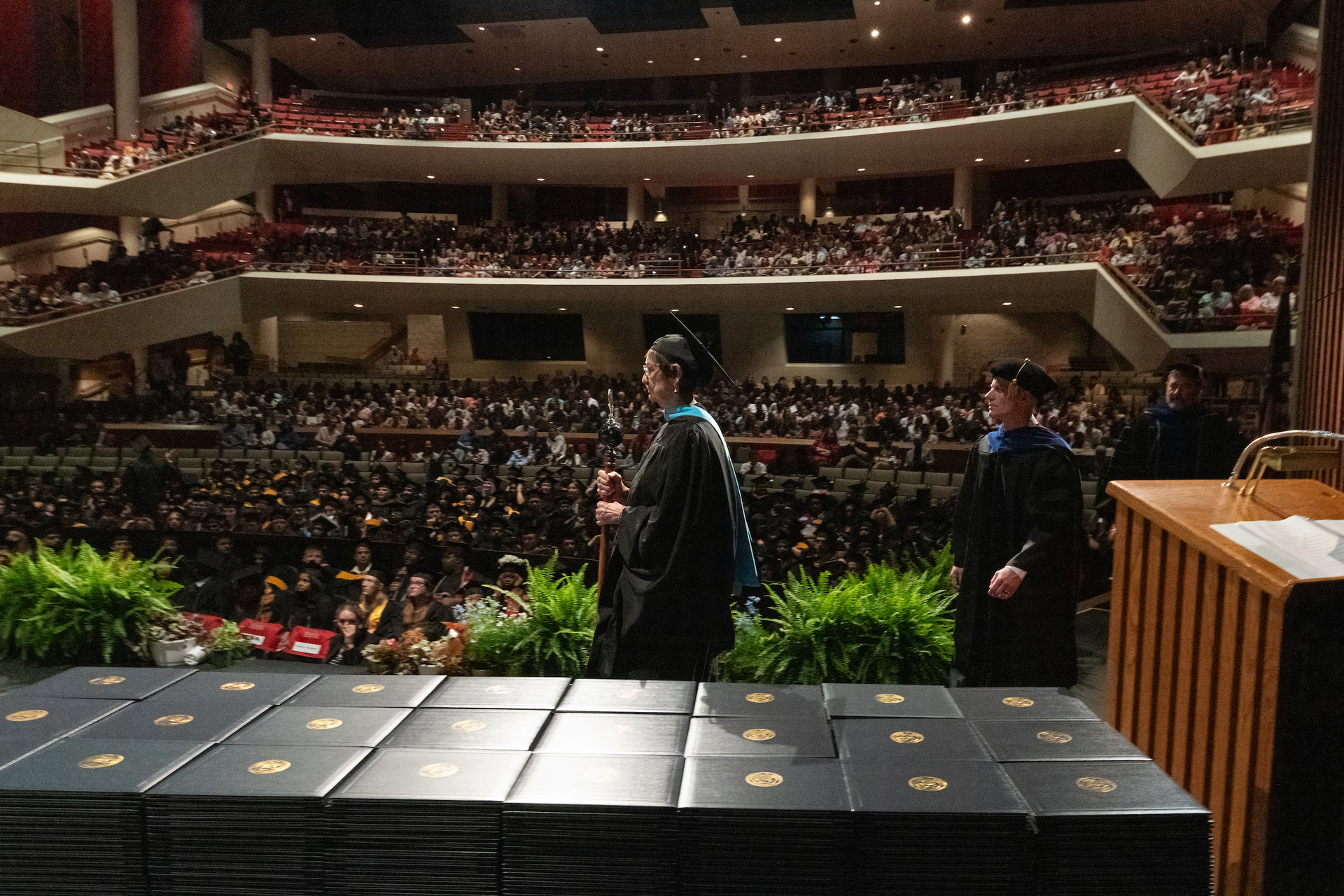 The student marshal, dressed in regalia, walks across the Sangamon Auditorium stage with the audience in the background.